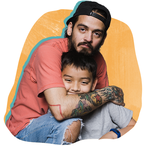 Hispanic man with backwards baseball cap, tattoos and a serious expression hugs a little boy who's smiling.