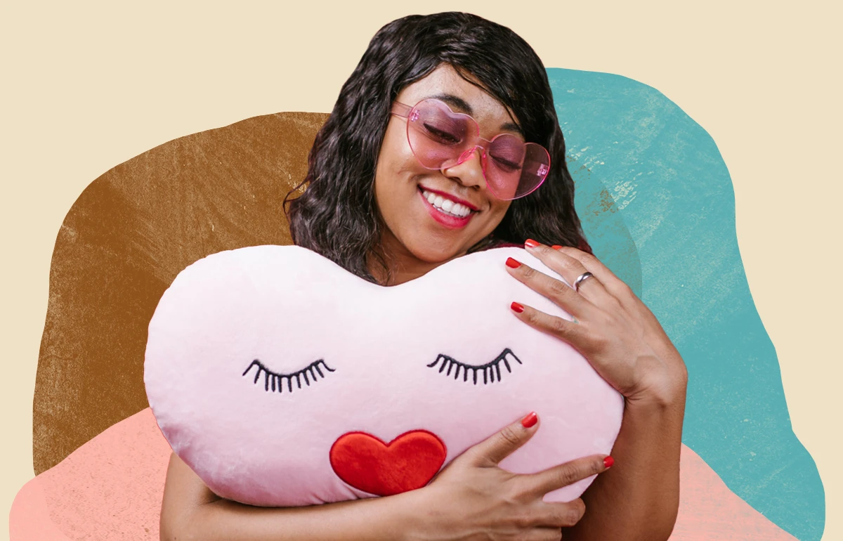 Smiling Black woman with heart-shaped glasses hugging a heart pillow