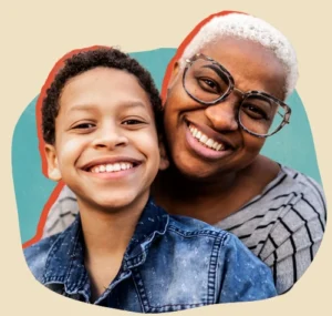 Black woman with short white hair and funky glasses smiling and holding a school-aged mixed-race boy in jean jacket, also smiling.