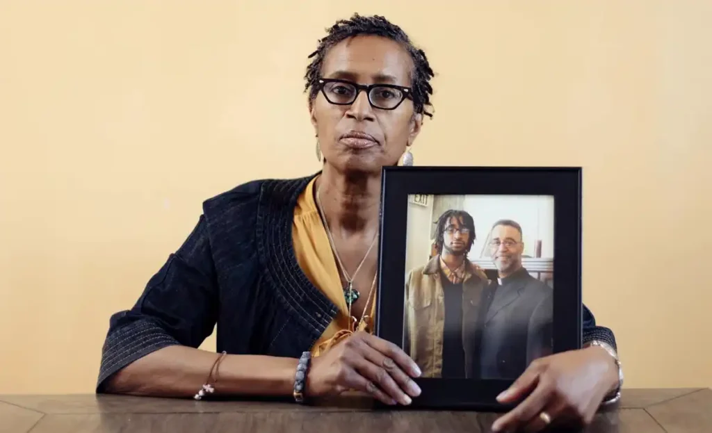 Black woman with short hair and serious expression holding a framed photo of two men