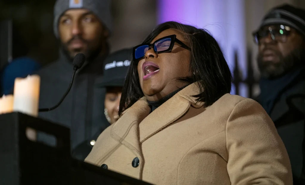 Black woman wearing a winter coat vocalizing passionately at a microphone at an evening vigil